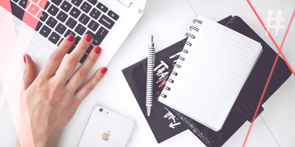 Mesa de escritório com notebook, caderno, caneta, celular com uma mão feminina digitando com esmalte vermelho nas unhas
